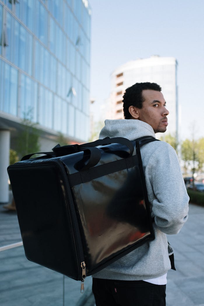 A courier in a city environment, carrying a large black delivery backpack outdoors.
